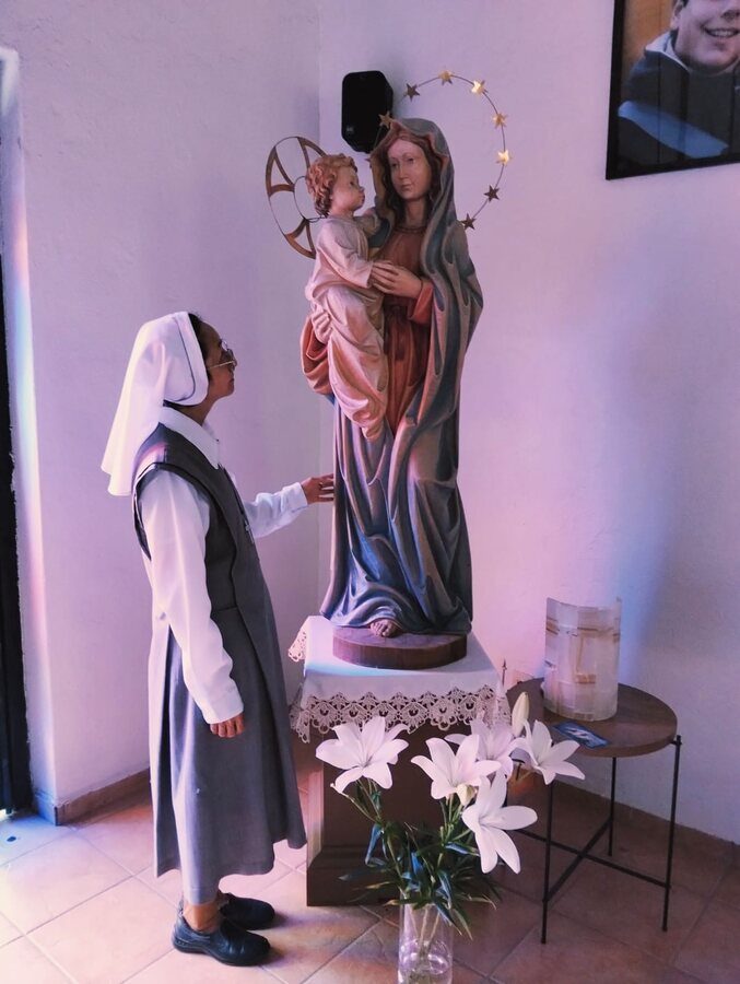 Una hermana contempla de pie una imagen de la Virgen con el Niño, junto a un ramo de azucenas.