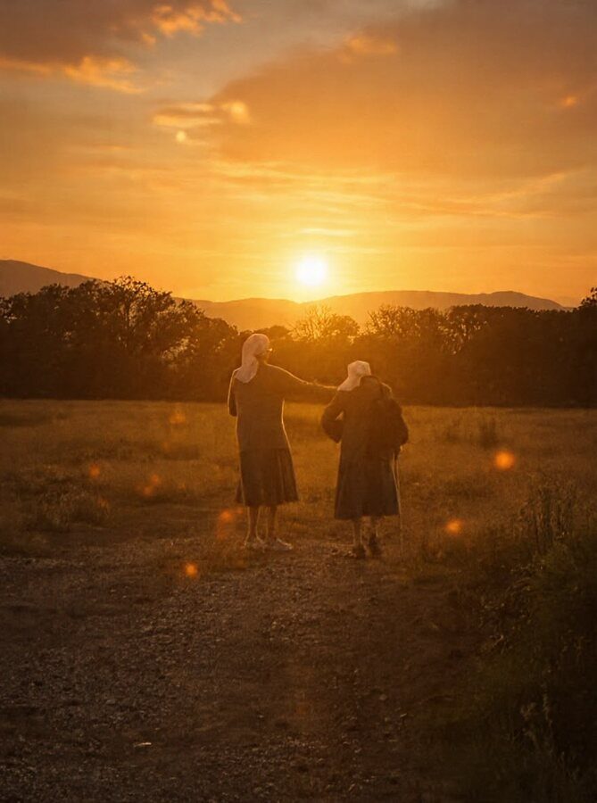 Dos hermanas caminan juntas por el campo al atardecer, con el sol poniéndose entre las montañas.