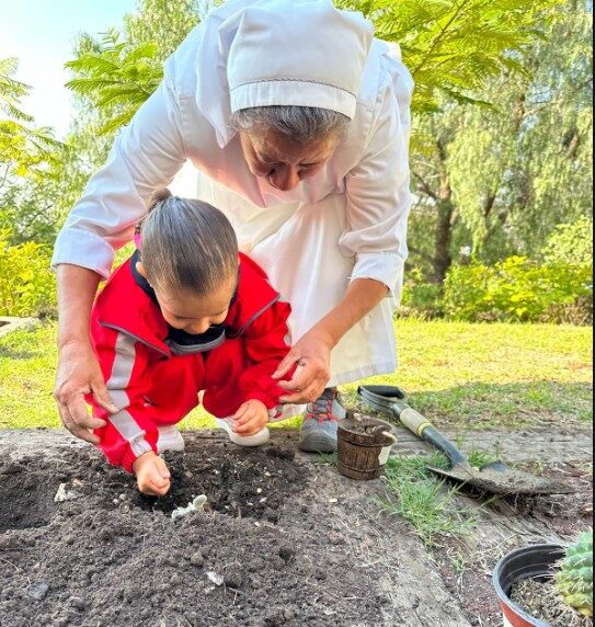 Una hermana marcelina acompaña a una niña que siembra con sus manos en la tierra de un huerto.