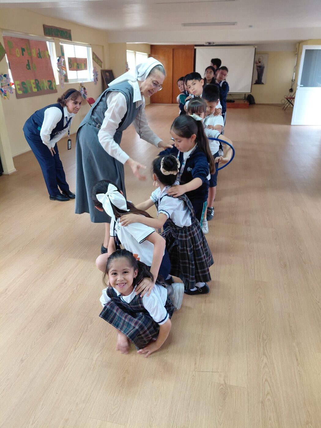 Una hermana marcelina juega con un grupo de alumnas y alumnos en el salón del colegio.